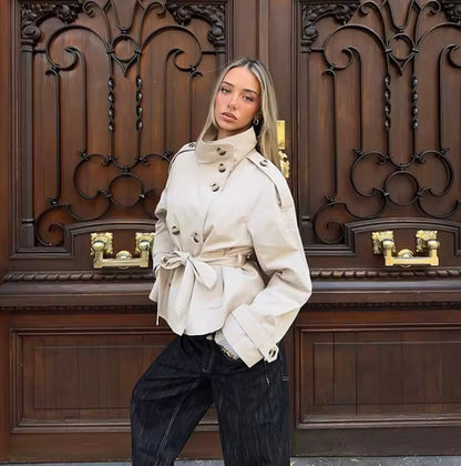 A woman posing in front of a large, ornate dark brown wooden door with gold hardware. She is wearing a light beige trench coat with a tie waist and dark charcoal gray wide-leg jeans.