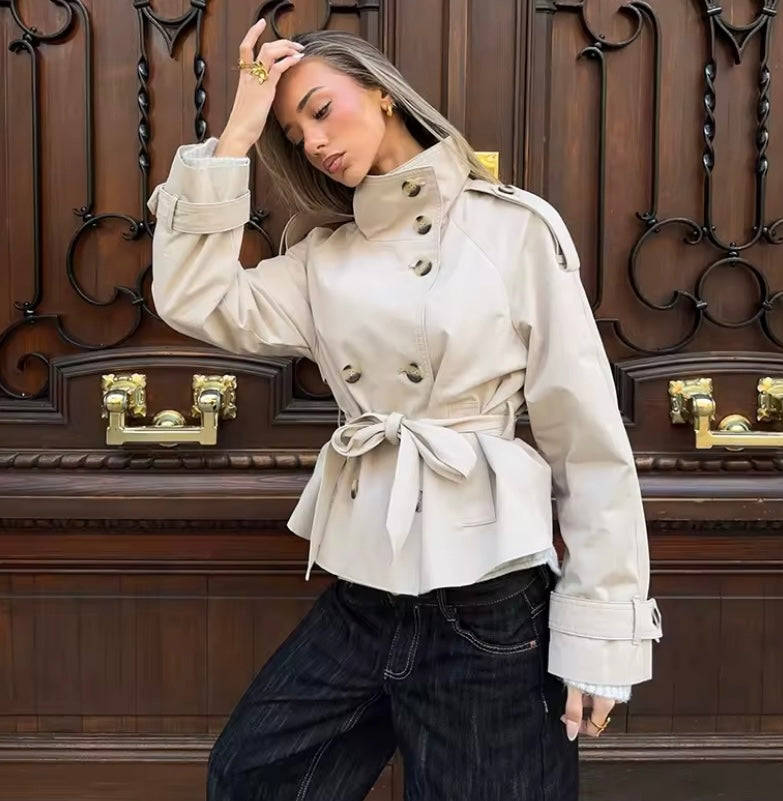 A woman models a beige double-breasted trench coat with a tie waist belt, paired with dark wide-leg jeans, posing in front of a large dark wood paneled door with gold handles.