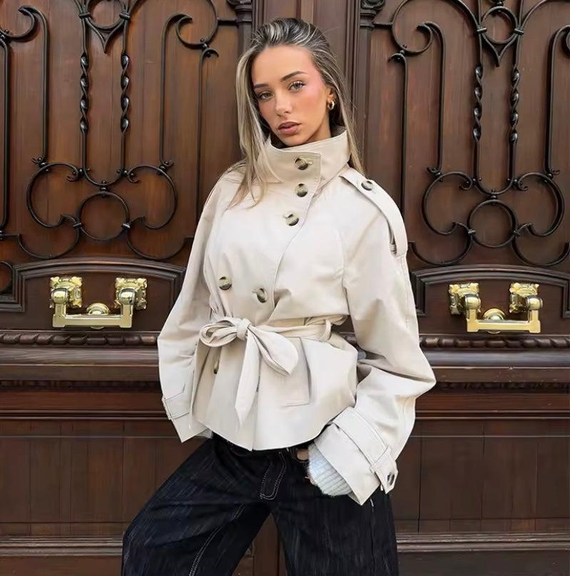 A woman models a beige double-breasted belted coat with wide sleeves, paired with dark wide-leg jeans, standing in front of a large ornate dark brown wooden door with gold hardware.