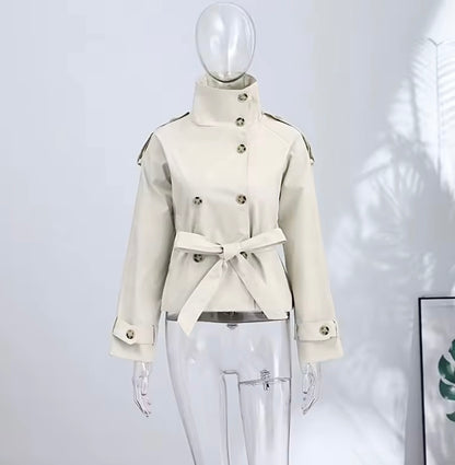 A cream-colored double-breasted jacket with a high stand-up collar and waist tie, displayed on a clear mannequin against a light gray wall.