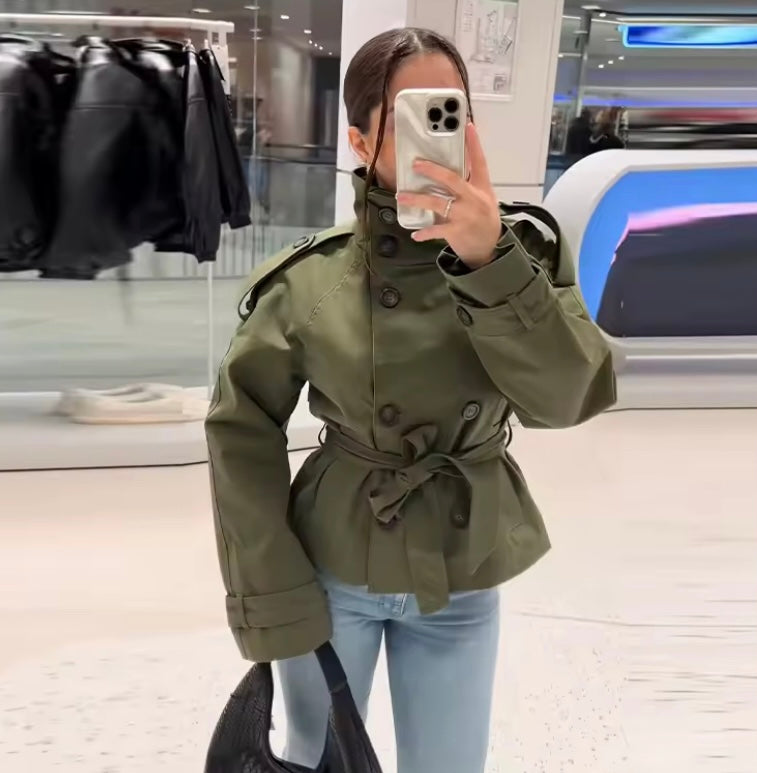 A woman takes a mirror selfie in a clothing store wearing an olive green belted trench-style jacket and light blue jeans.