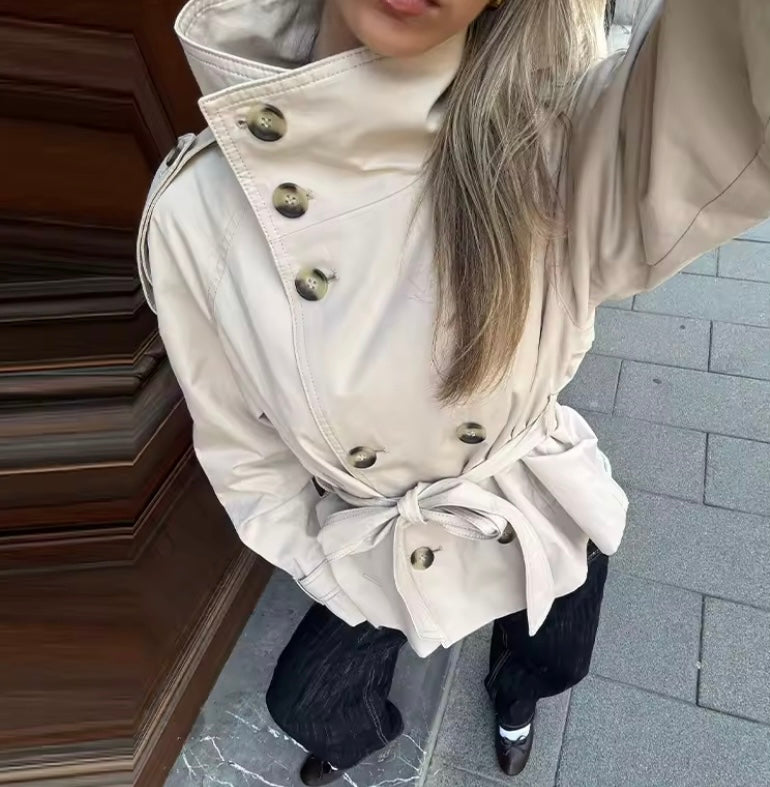 A person wearing a light beige double-breasted trench coat with a tie waist belt, paired with black wide-leg trousers and black shoes, standing on a gray paved surface next to a dark wooden door.