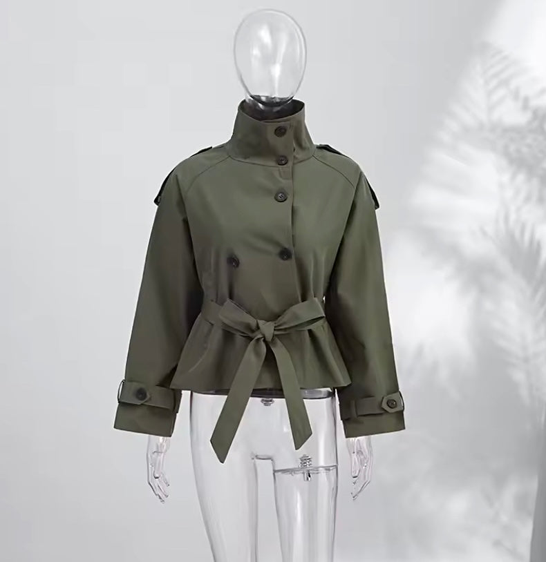 An olive green military-style jacket with a high stand collar, front buttons, shoulder epaulets, and a matching waist tie, displayed on a clear mannequin against a light gray background with soft leaf shadows.