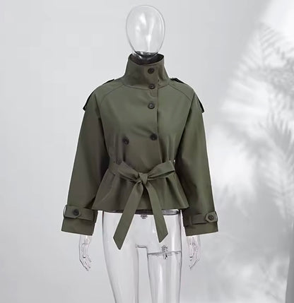 An olive green military-style jacket with a high stand collar, front buttons, shoulder epaulets, and a matching waist tie, displayed on a clear mannequin against a light gray background with soft leaf shadows.